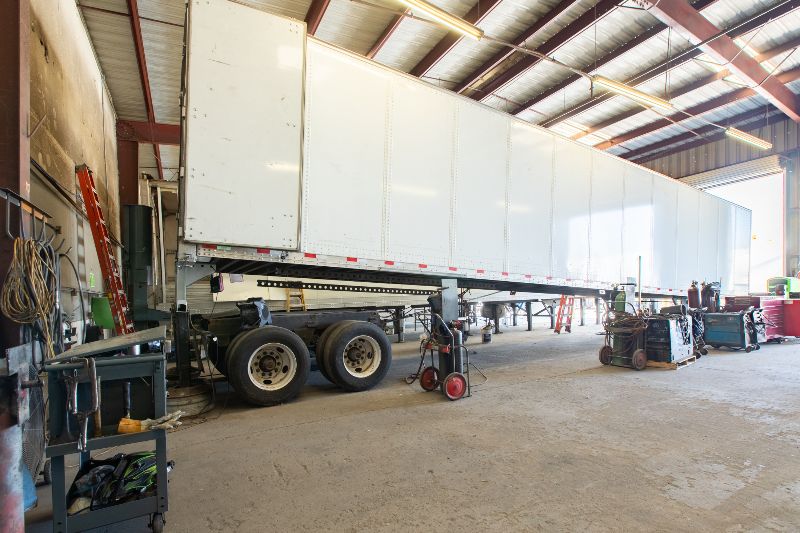 Large semi-trailer inside a repair shop, elevated for maintenance, surrounded by tools, welding equipment, and ladders under bright overhead lighting.