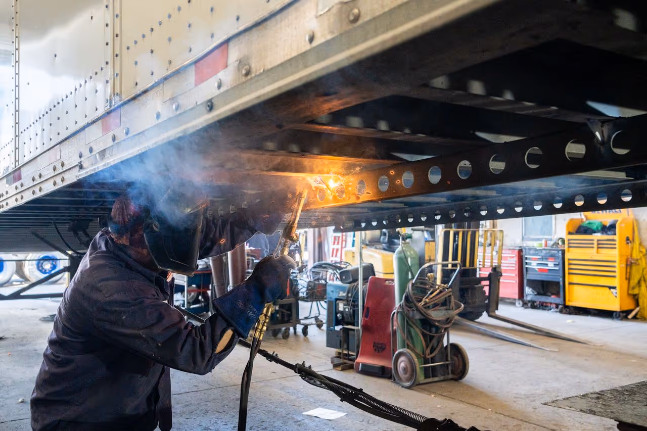 Technician welding structural beams beneath a trailer inside a repair facility, with sparks flying and shop equipment visible in the background.