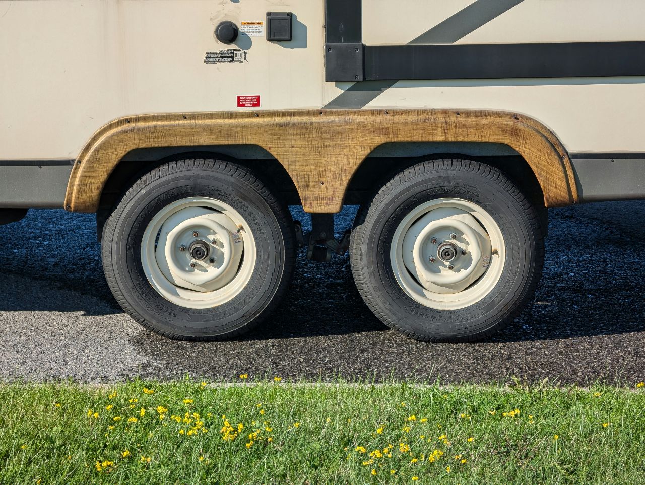 Close-up of a tandem axle trailer’s dual wheels with white rims, parked on asphalt beside grass and small yellow flowers.