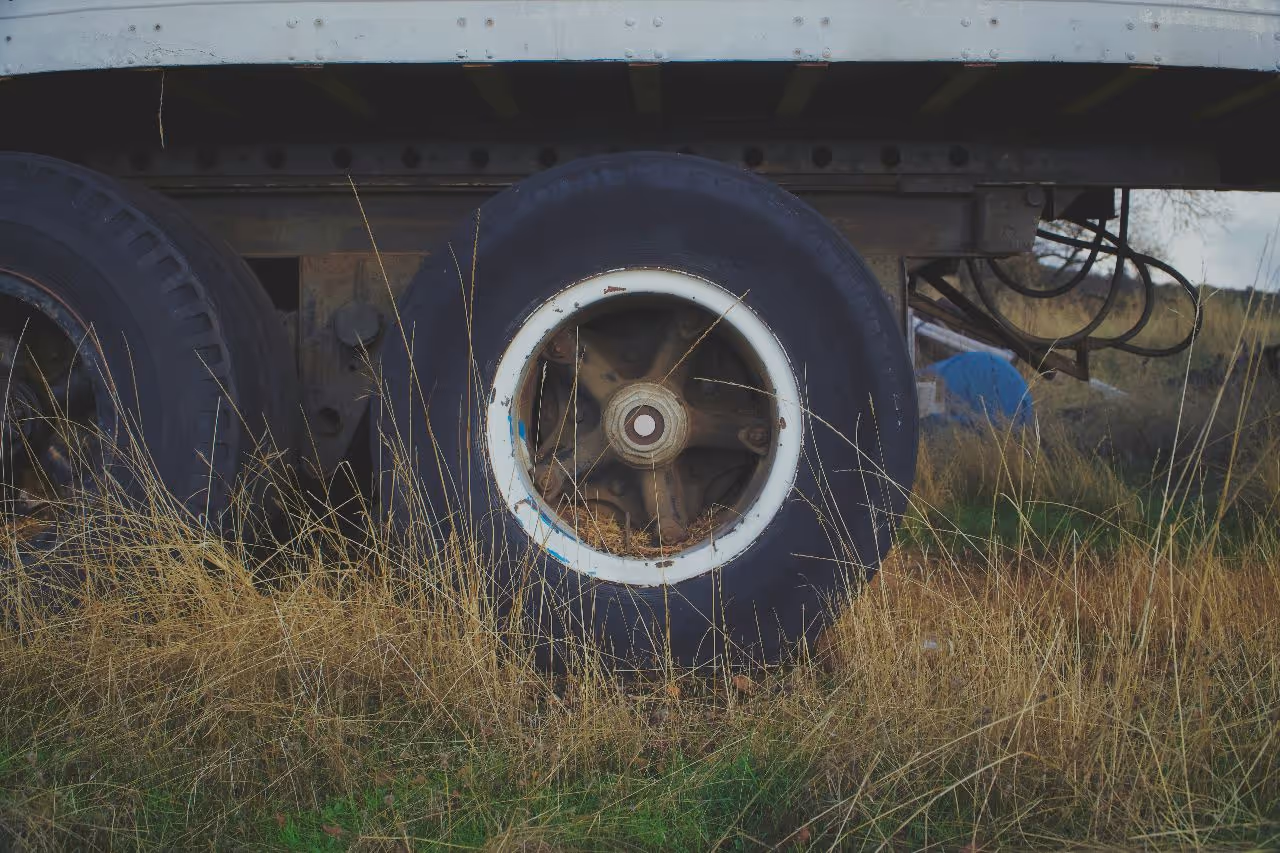 Dual semi-trailer wheels partially hidden by tall grass, highlighting weathered tires and metal undercarriage in rural outdoor setting.