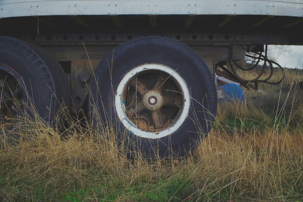 Dual semi-trailer wheels partially hidden by tall grass, highlighting weathered tires and metal undercarriage in rural outdoor setting.