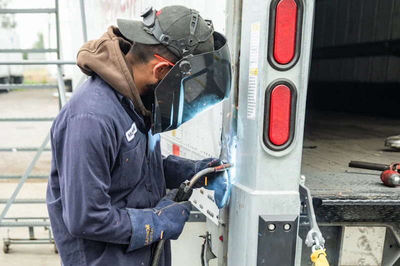Trailer fabrication showing technician welding metal panel on truck trailer with sparks and visible red taillights.