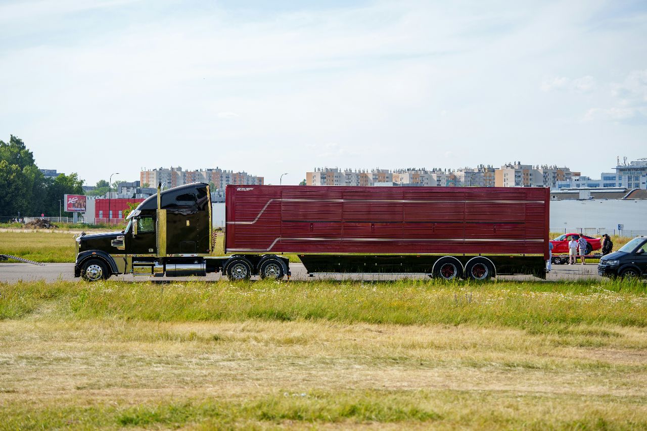 Black semi-truck hauling a large red trailer across an open roadway with grassy fields and buildings in the background.
