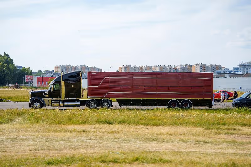 Black semi-truck hauling a large red trailer across an open roadway with grassy fields and buildings in the background.