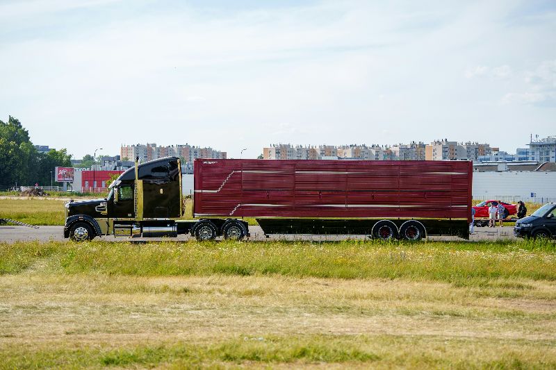 Black semi-truck hauling a large red trailer across an open roadway with grassy fields and buildings in the background.