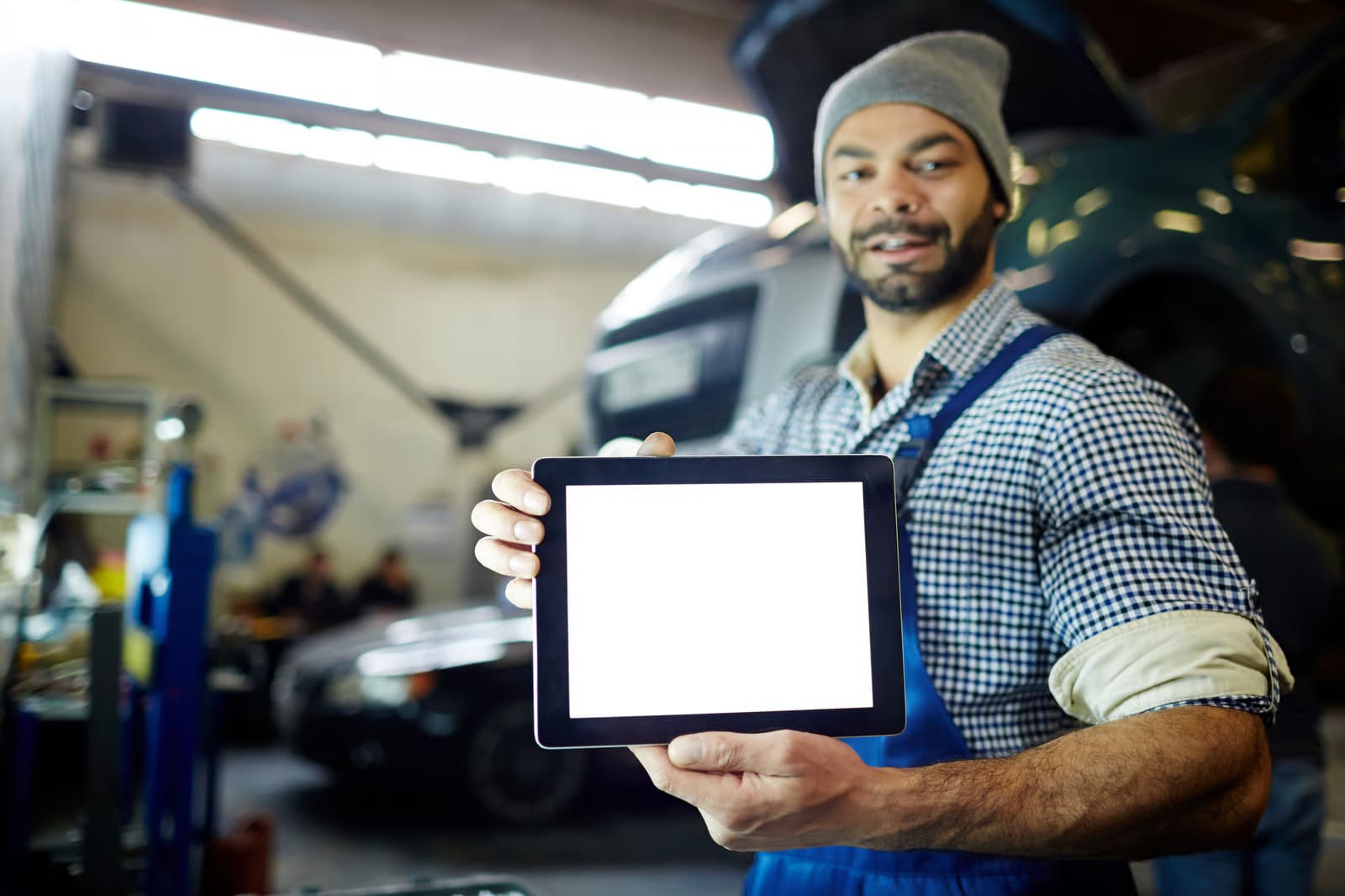 A smiling auto mechanic in overalls holding up a digital tablet with a blank white screen inside a car repair shop.
