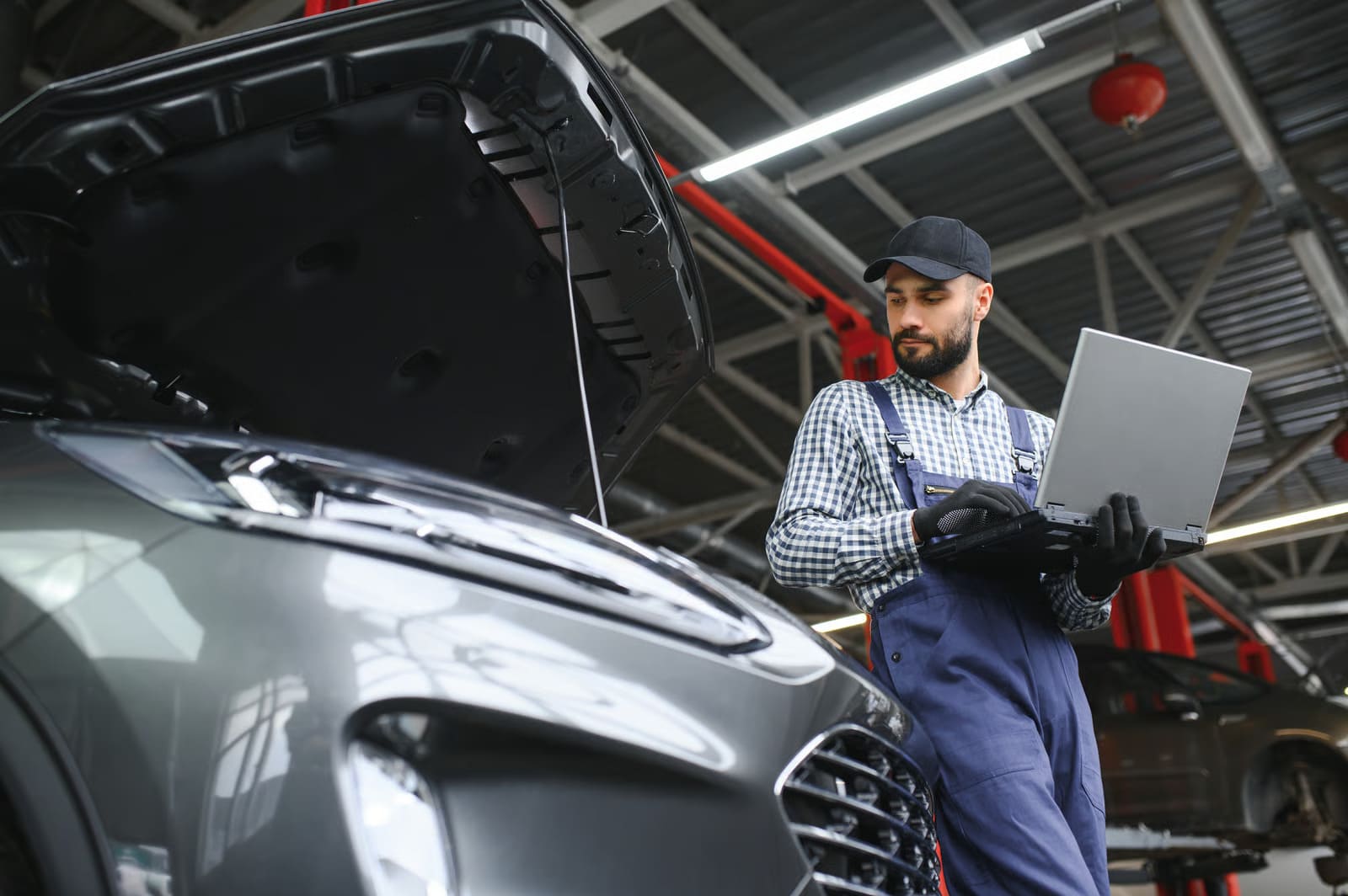 Professional car mechanic using a diagnostic laptop next to an open vehicle hood in an auto repair shop.