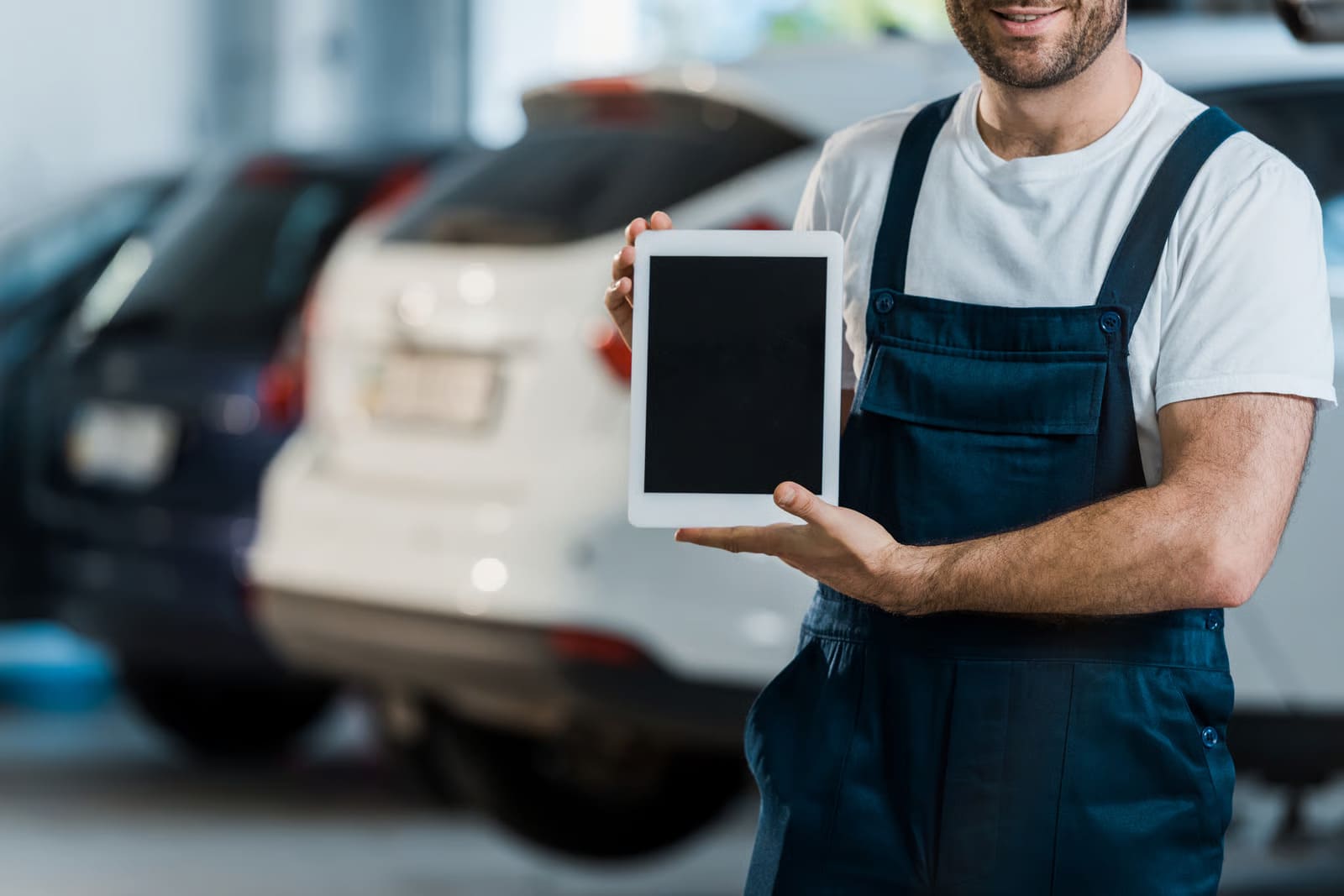 Auto mechanic in dark blue overalls holding up a blank digital tablet in an auto repair workshop with cars parked in the background.