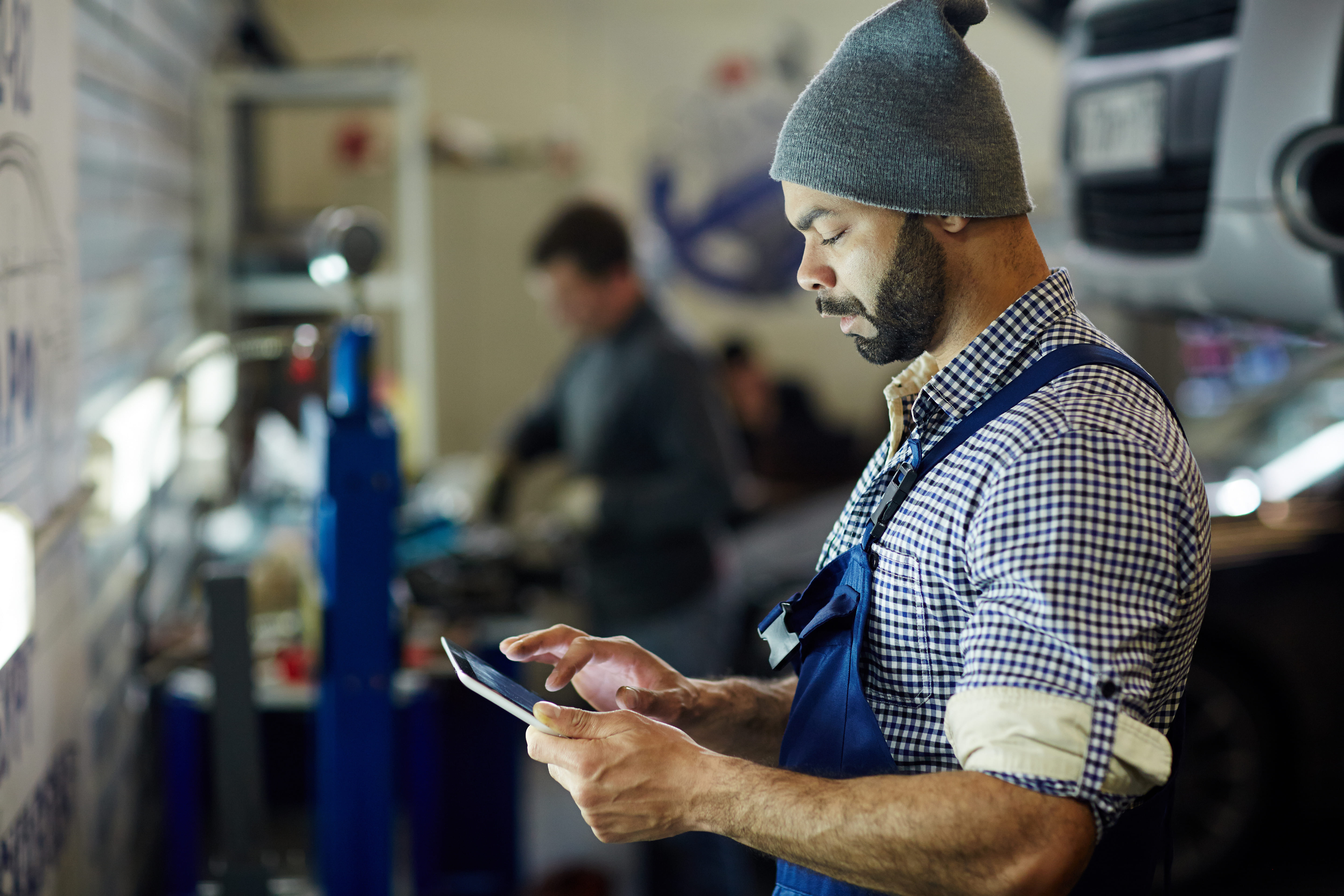 Automotive technician in blue overalls and a knit beanie using a digital tablet inside a busy auto repair workshop with vehicles on hydraulic lifts.