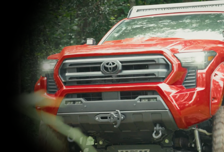 Front view of a red Toyota truck driving through a forested area with headlights on.