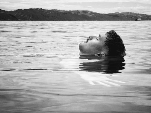 Black and white photo of a man floating calmly on water with his eyes closed and a mountainous shoreline in the background.