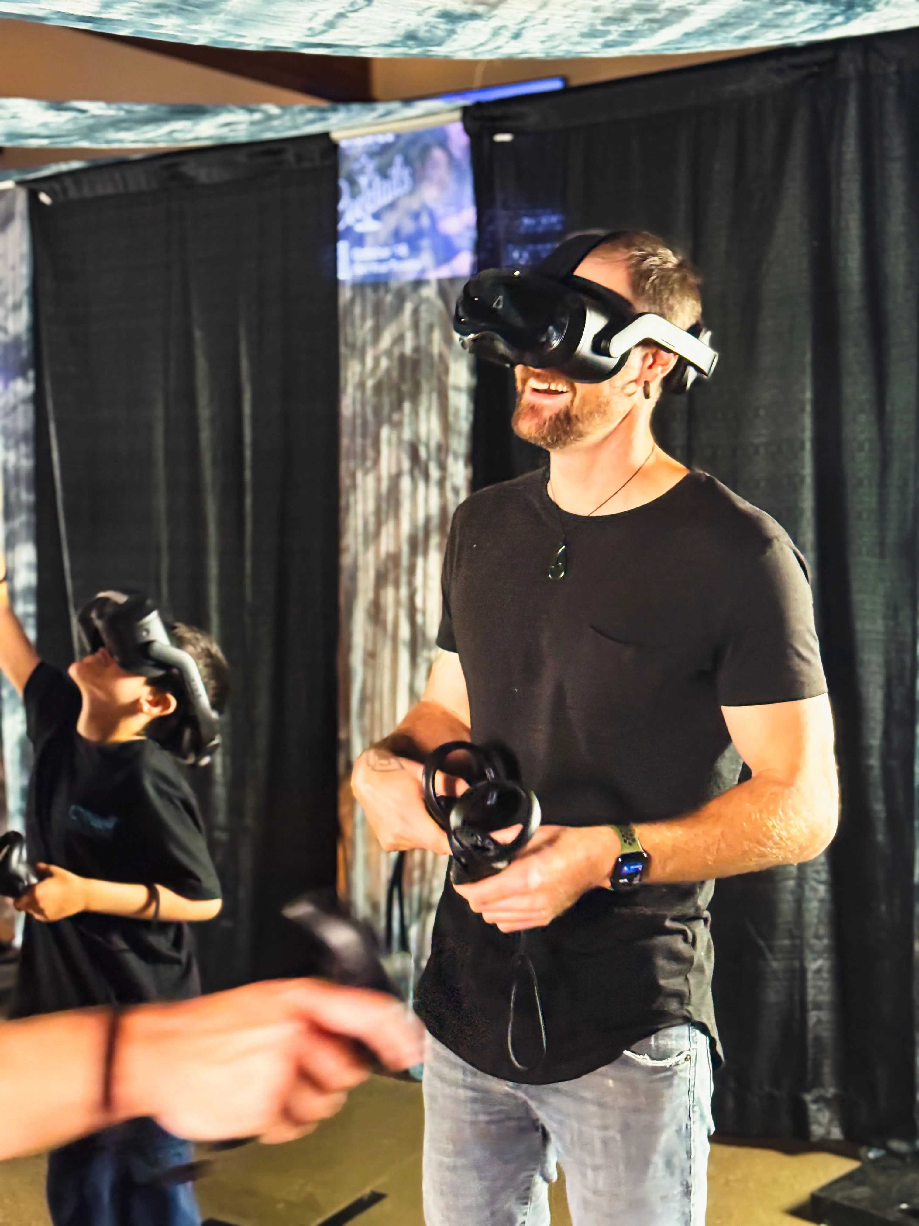An adult guest smiles broadly inside a VR headset while a child plays beside him.
