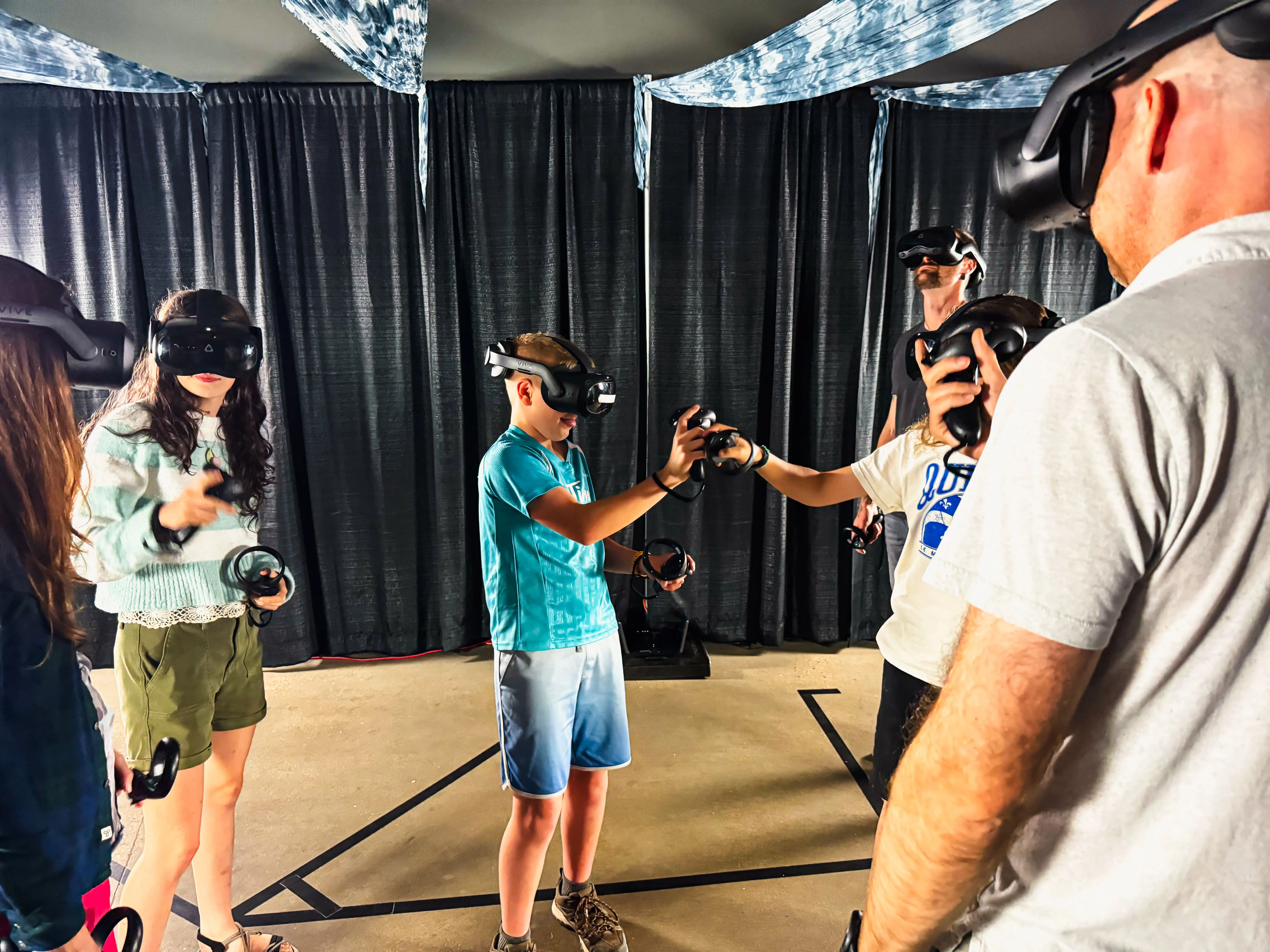 A group of guests in VR headsets reach toward the same point in the shared virtual world.