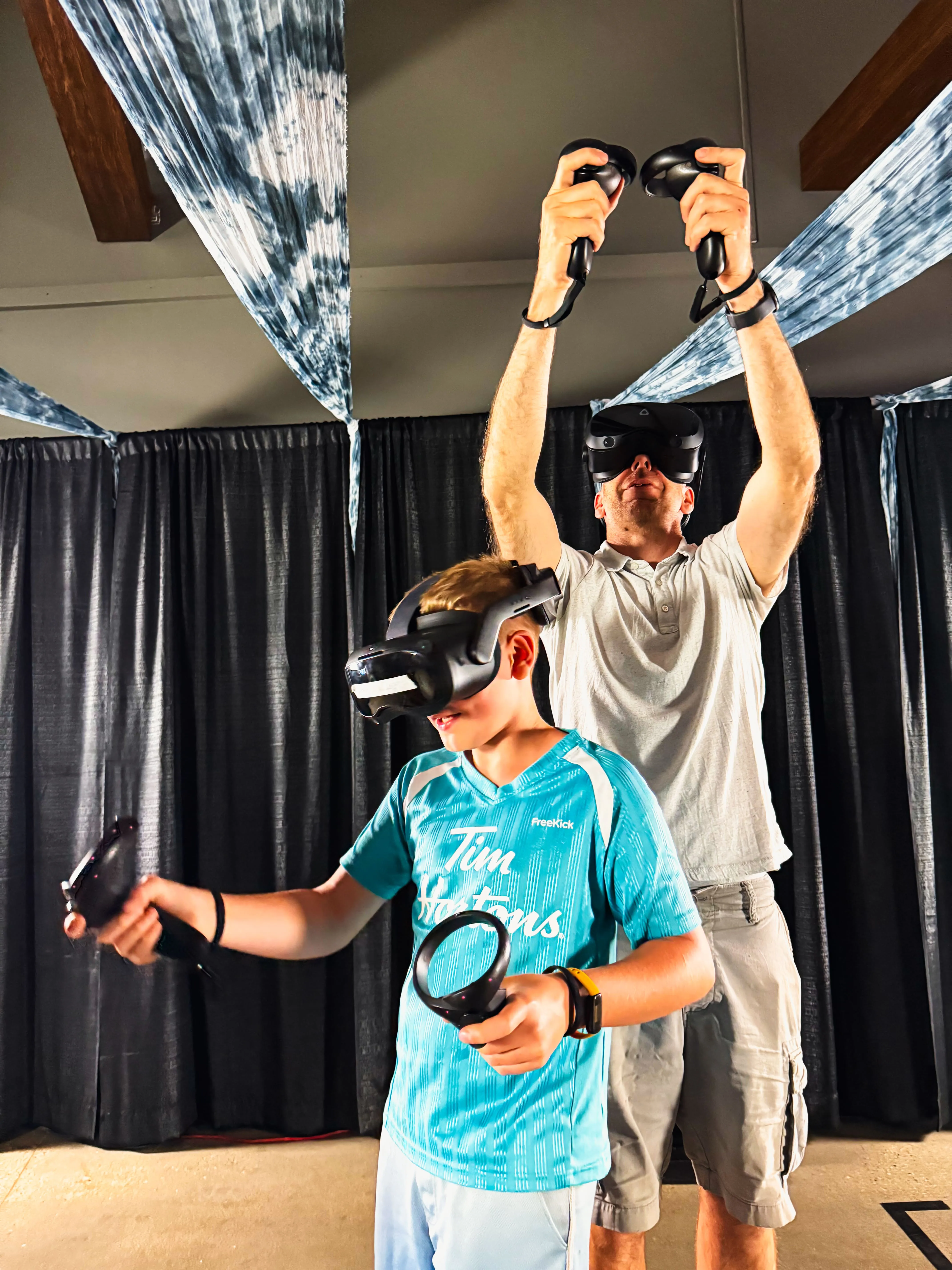 An adult guest raises both controllers overhead beside a boy in a VR headset during a shared session.