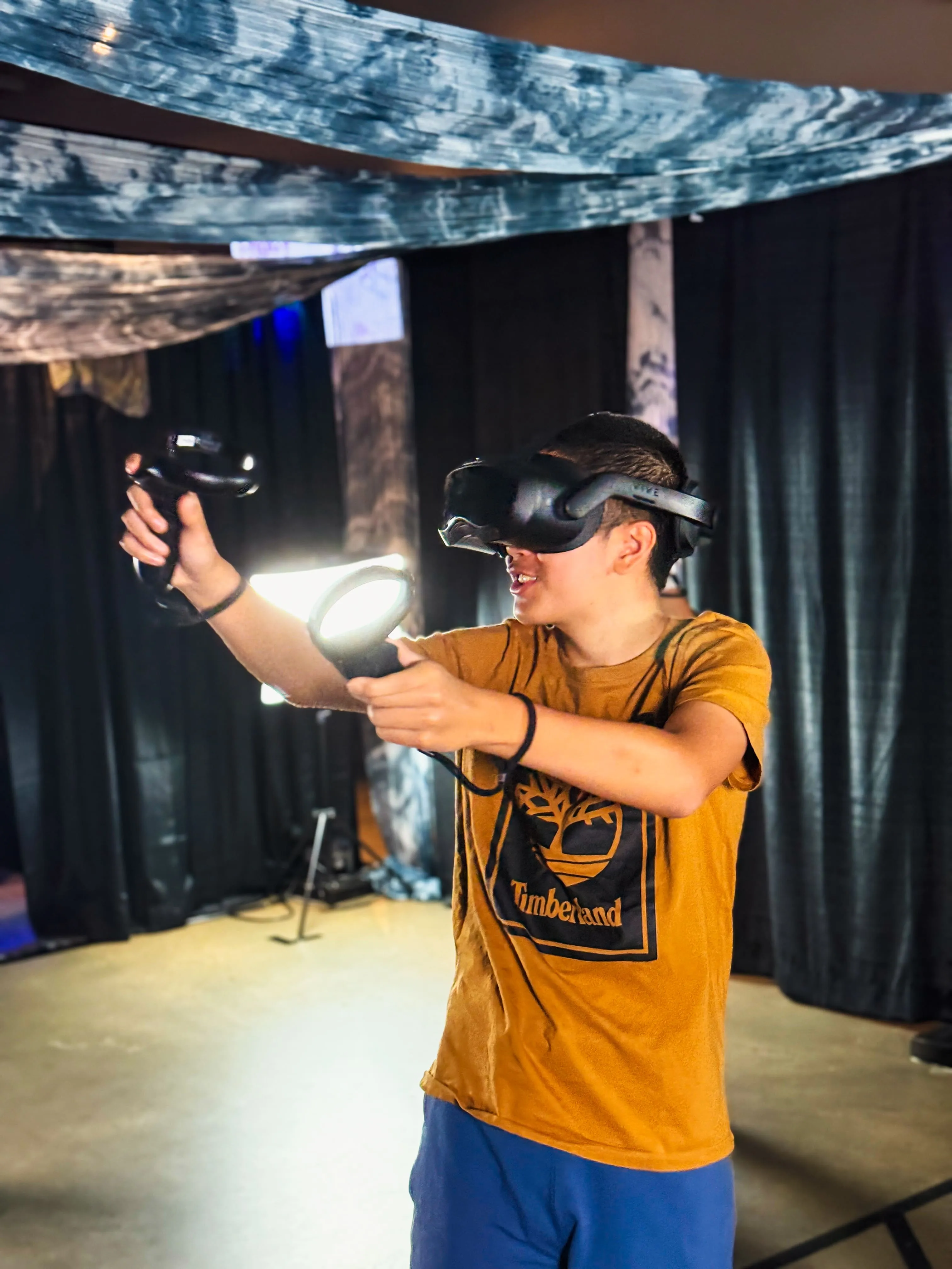 A teenage guest in a VR headset reaches out with both controllers, smiling mid-session.