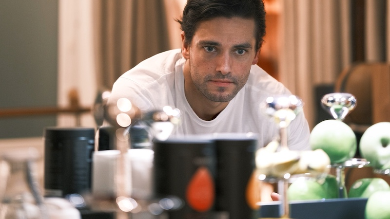 Un homme en chemise blanche regarde attentivement plusieurs pommes vertes et divers objets posés sur une table.