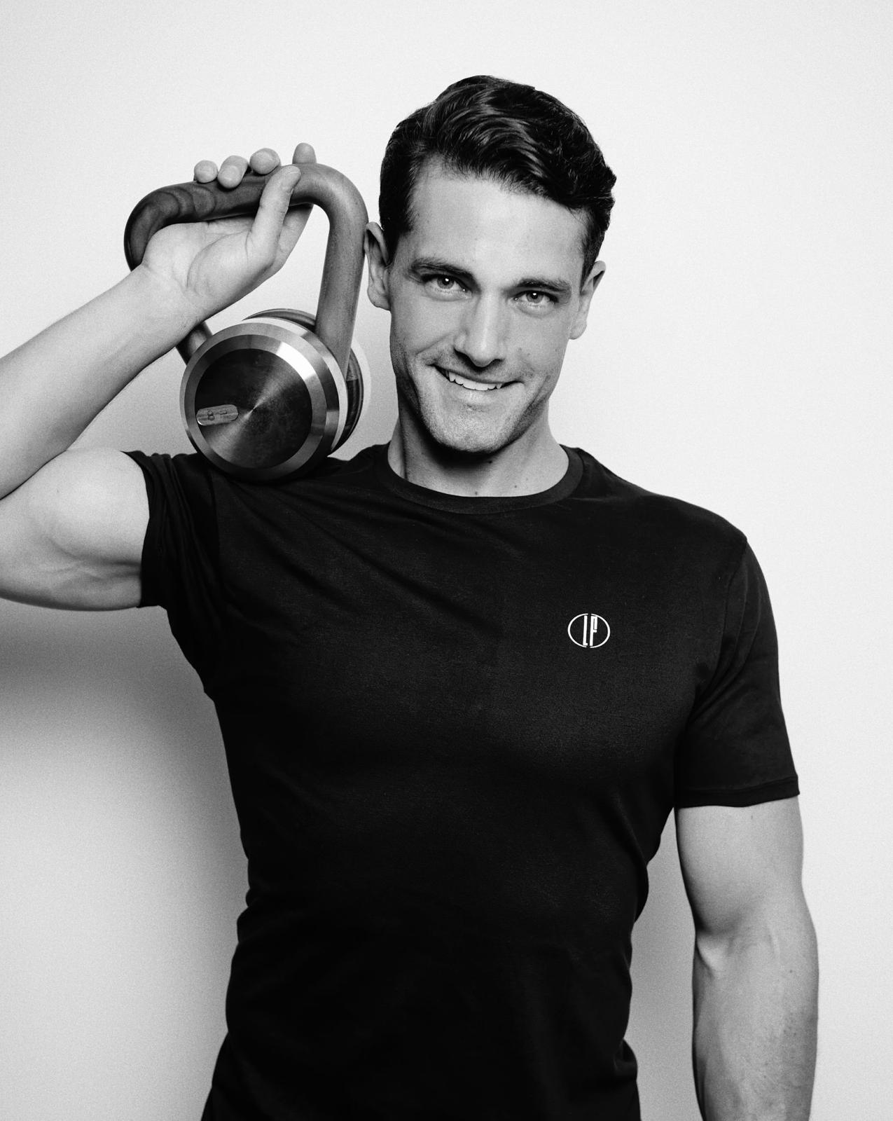 Louis Fabre, entrenador sonriente, con una camiseta negra y una pesa rusa sobre el hombro izquierdo.