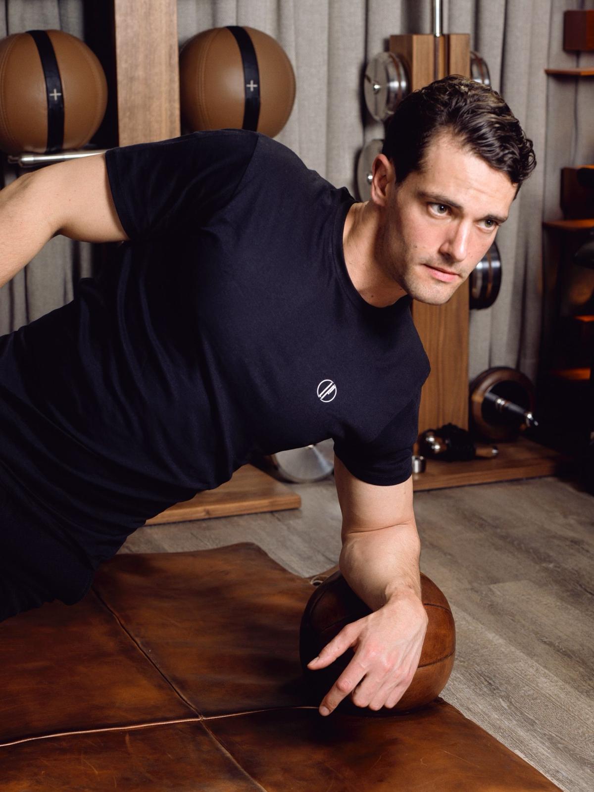 Louis Fabre Coaching con camiseta negra haciendo un ejercicio de abdominales laterales sobre un balón medicinal de cuero en un gimnasio.