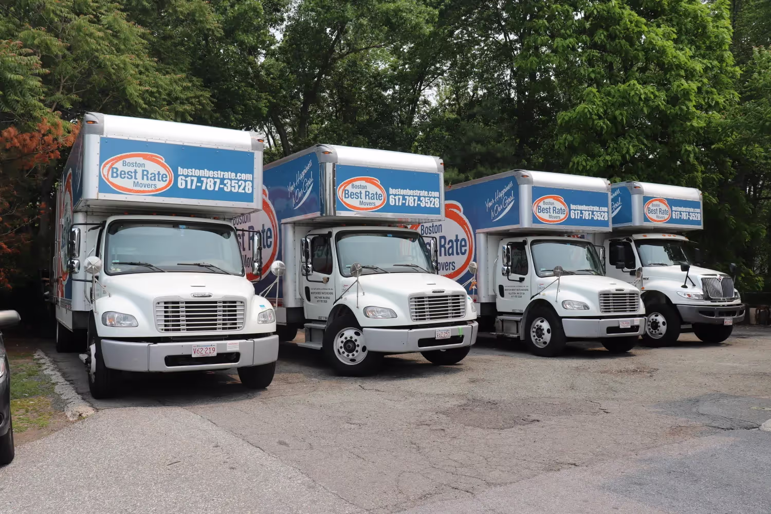 Four white moving trucks with blue and orange Boston Best Rate Movers signage parked in a row on a paved surface with trees in the background.