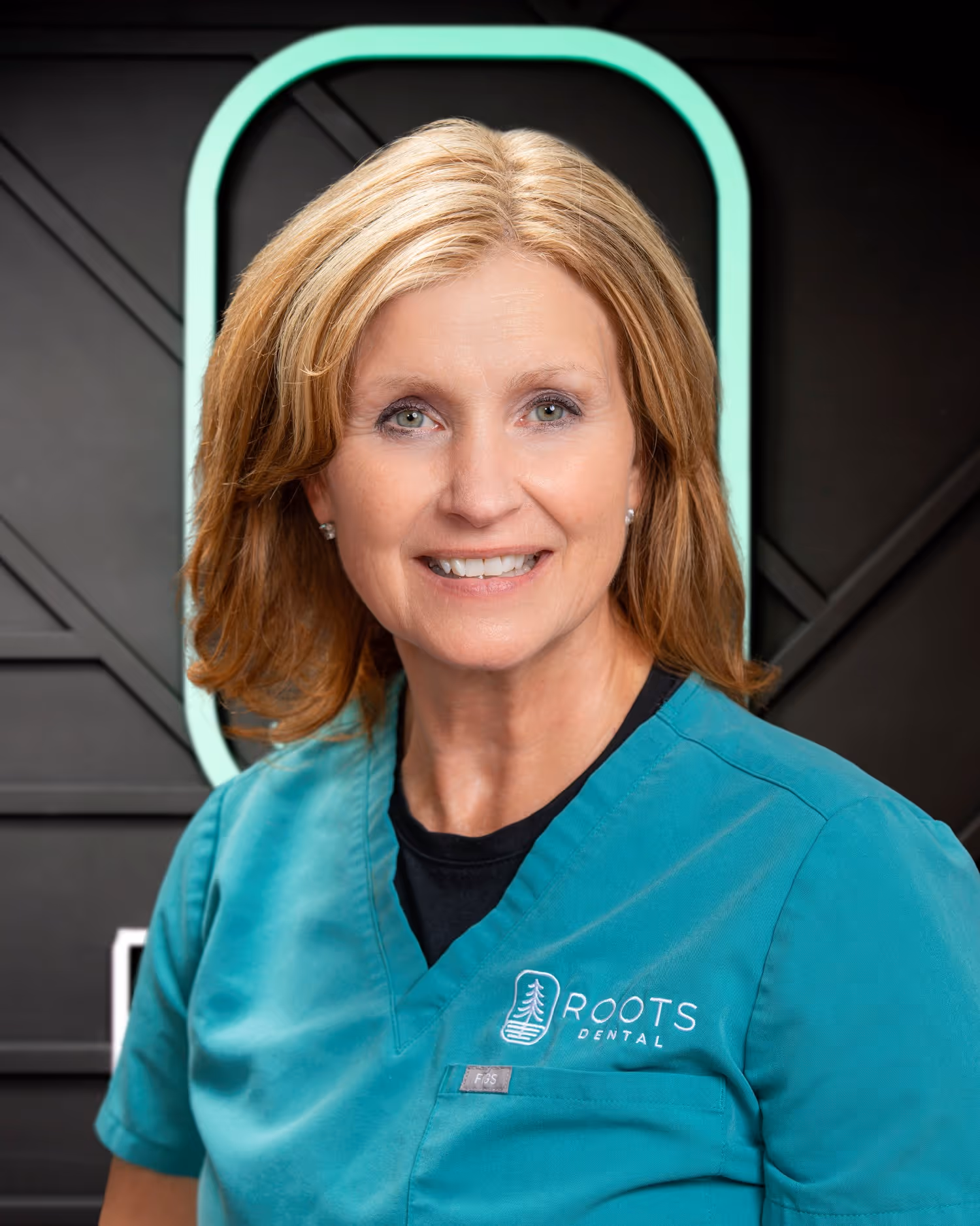 Smiling woman in teal Roots Dental scrubs with light brown hair and green eyes, standing against a dark background with a glowing green oval shape.