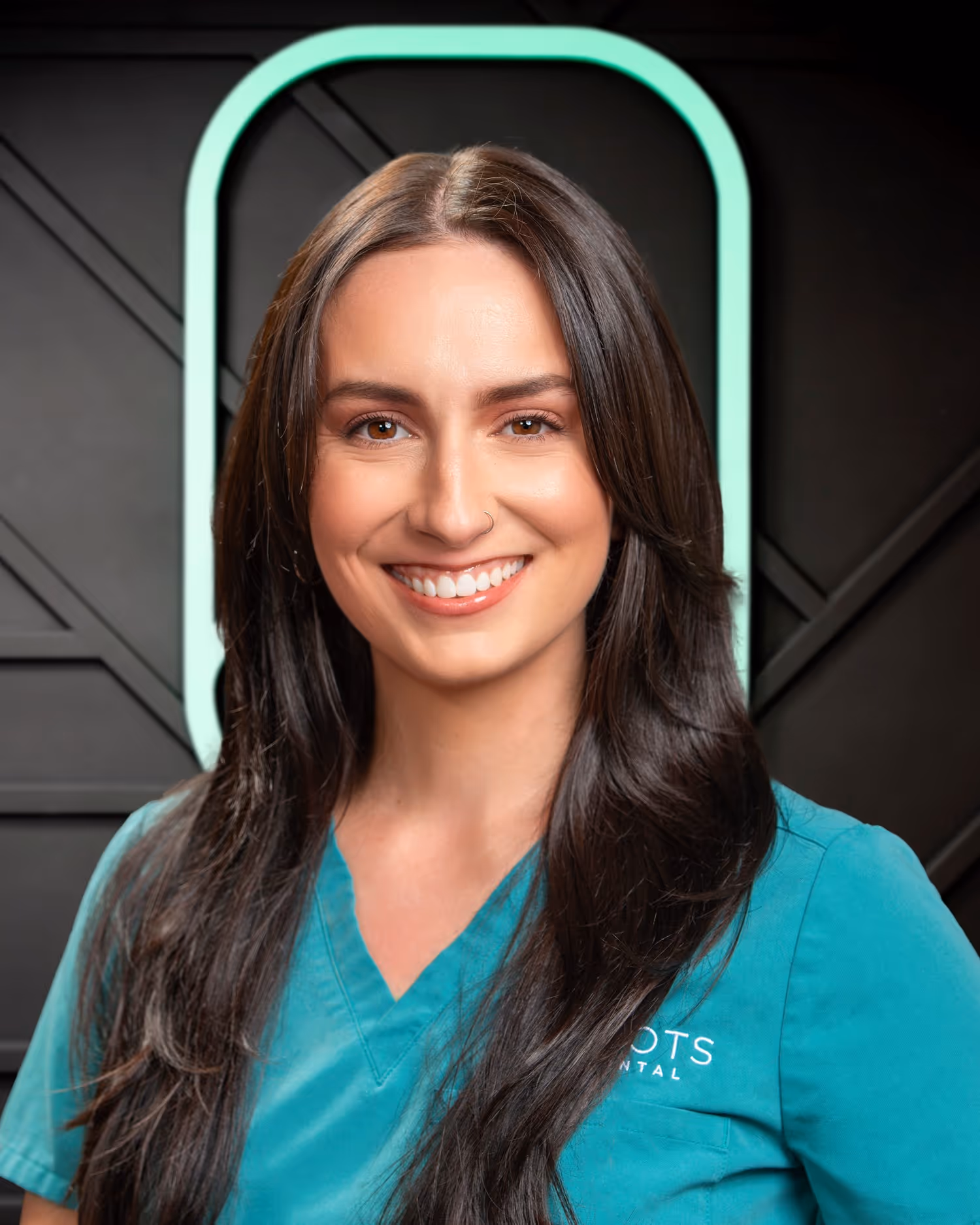 Smiling woman with long dark hair wearing teal scrubs standing in front of a dark wall with a mint-green neon arch.