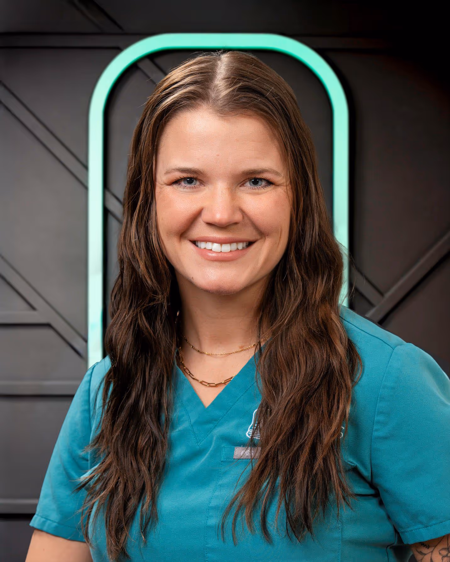 Smiling woman with long brown hair wearing teal medical scrubs and gold necklaces in front of a dark wall with a glowing green arch.