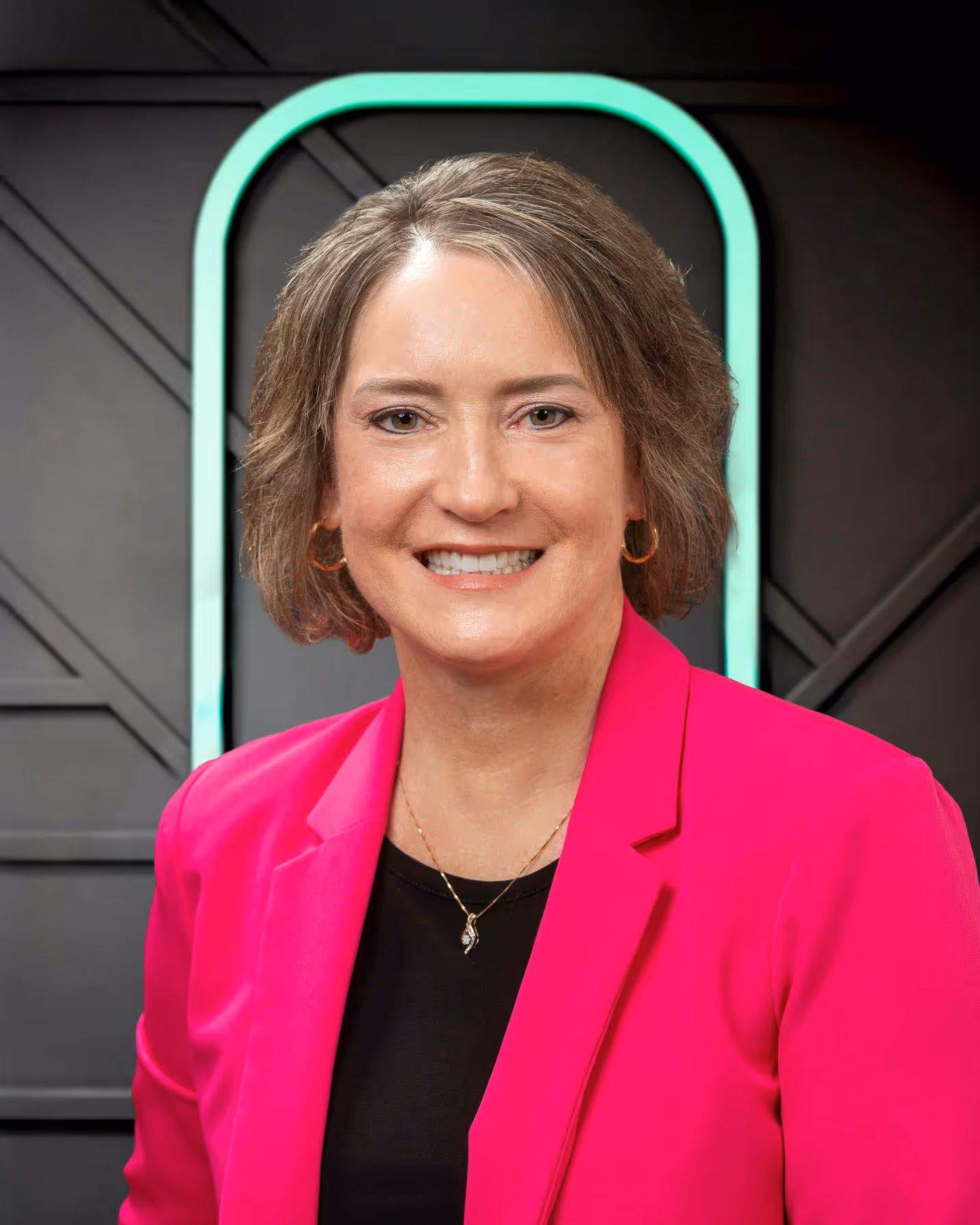 Smiling woman with short brown hair wearing a bright pink blazer, black top, and gold necklace against a dark background with a neon green arch.