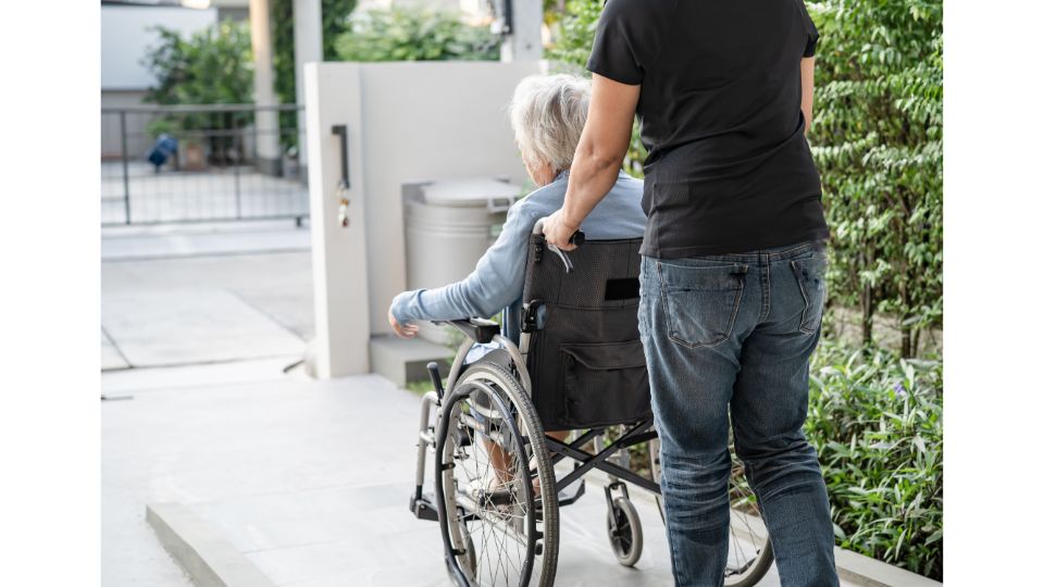 Pflegekraft unterstützt eine ältere pflegebedürftige Person im Rollstuhl und schiebt sie vorwärts.