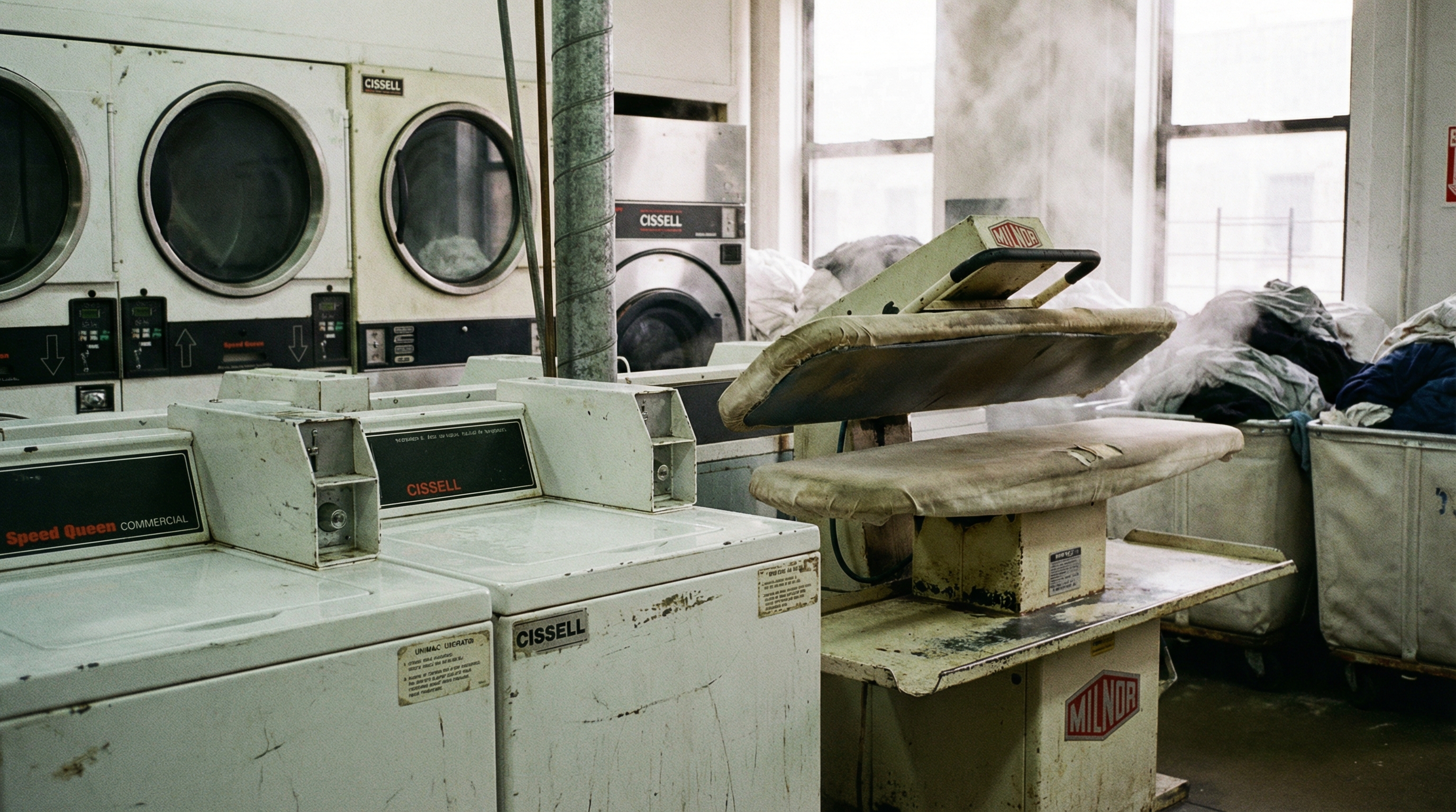 Close-up of various commercial laundry equipment including washers, dryers, and pressing machines with visible brand labels and condition details