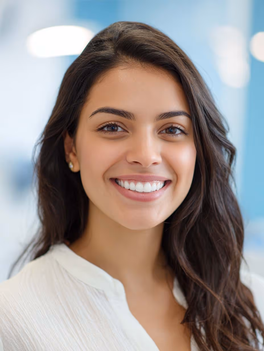 Jeune femme souriante aux cheveux bruns portant un chemisier blanc sur fond flou bleu clair.