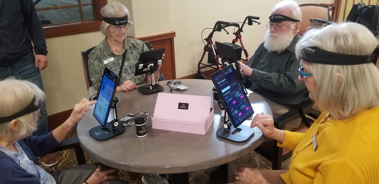 Elderly people, each wearing a Thinkie sensor, gather around table with tablets during technology demonstration.