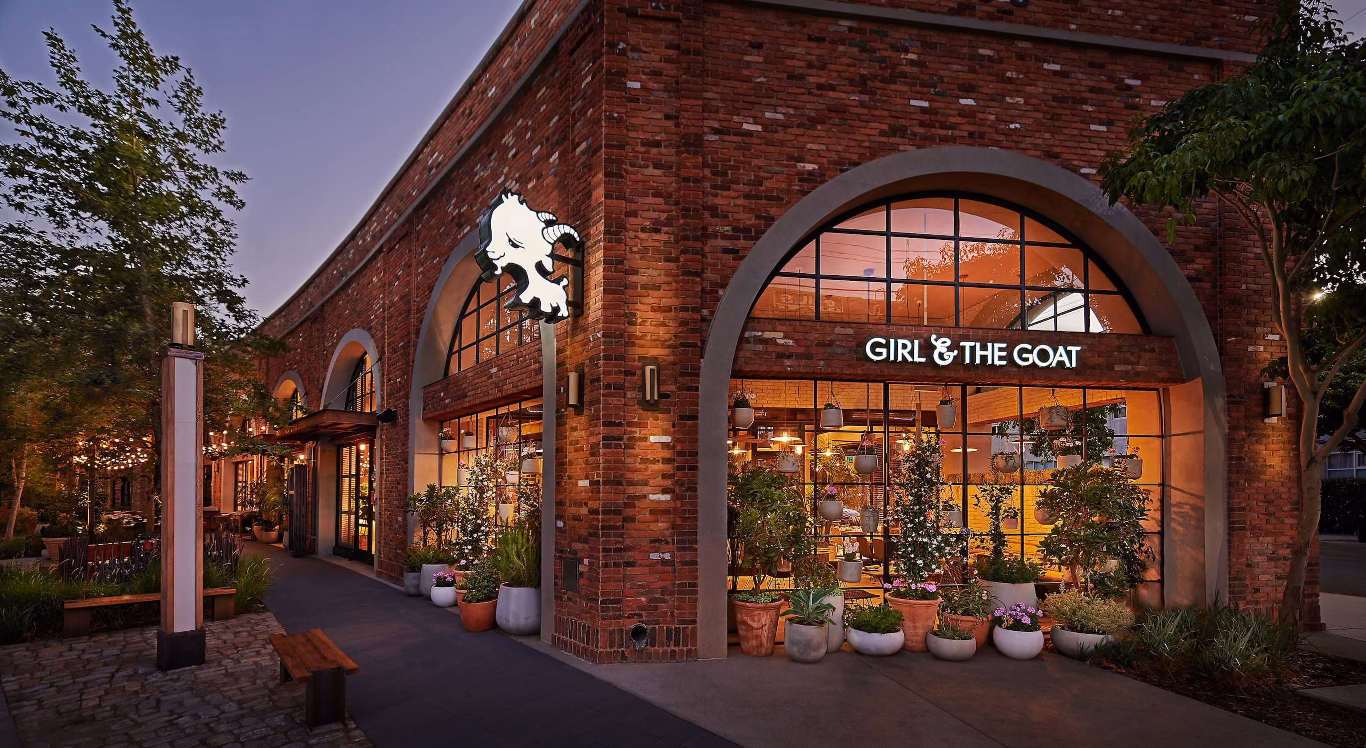 Brick restaurant exterior with arched windows, potted plants, and lit sign saying Girl & The Goat.