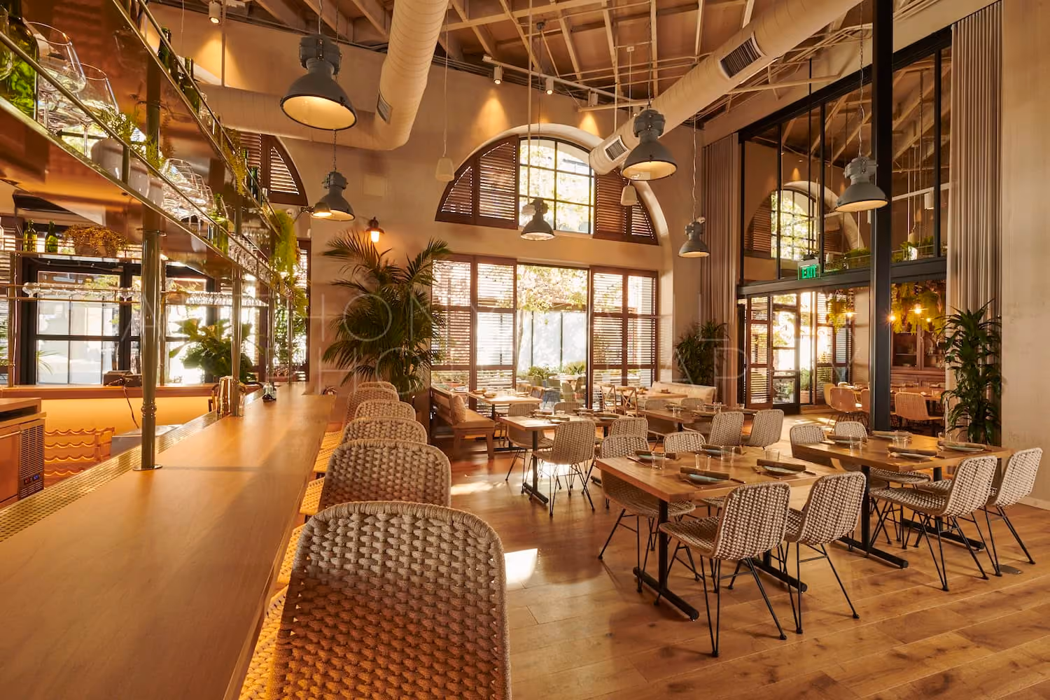 Cozy restaurant interior with wooden floors, wicker chairs, large windows, and industrial ceiling lights.