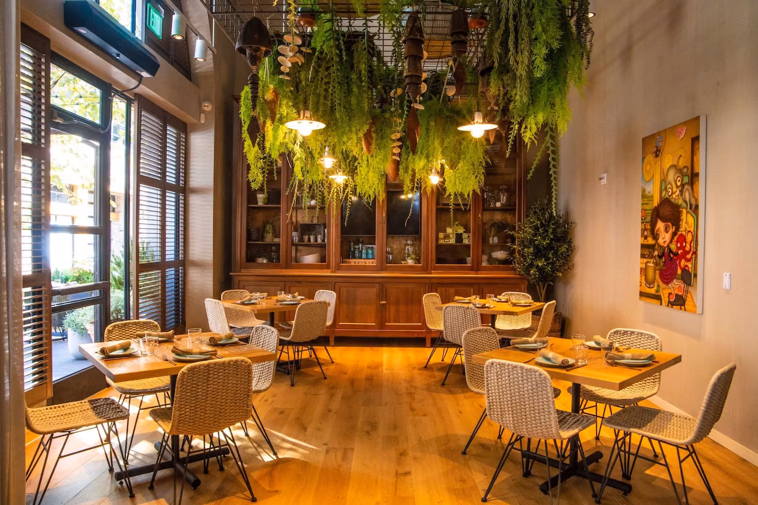 Cozy restaurant dining area with wooden tables, wicker chairs, hanging plants, and warm lighting.