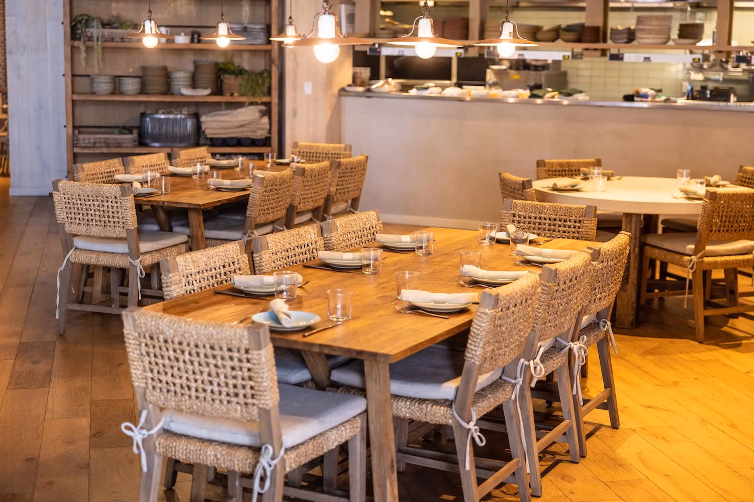 Warm restaurant dining area with wooden tables and woven chairs set with plates and glasses.
