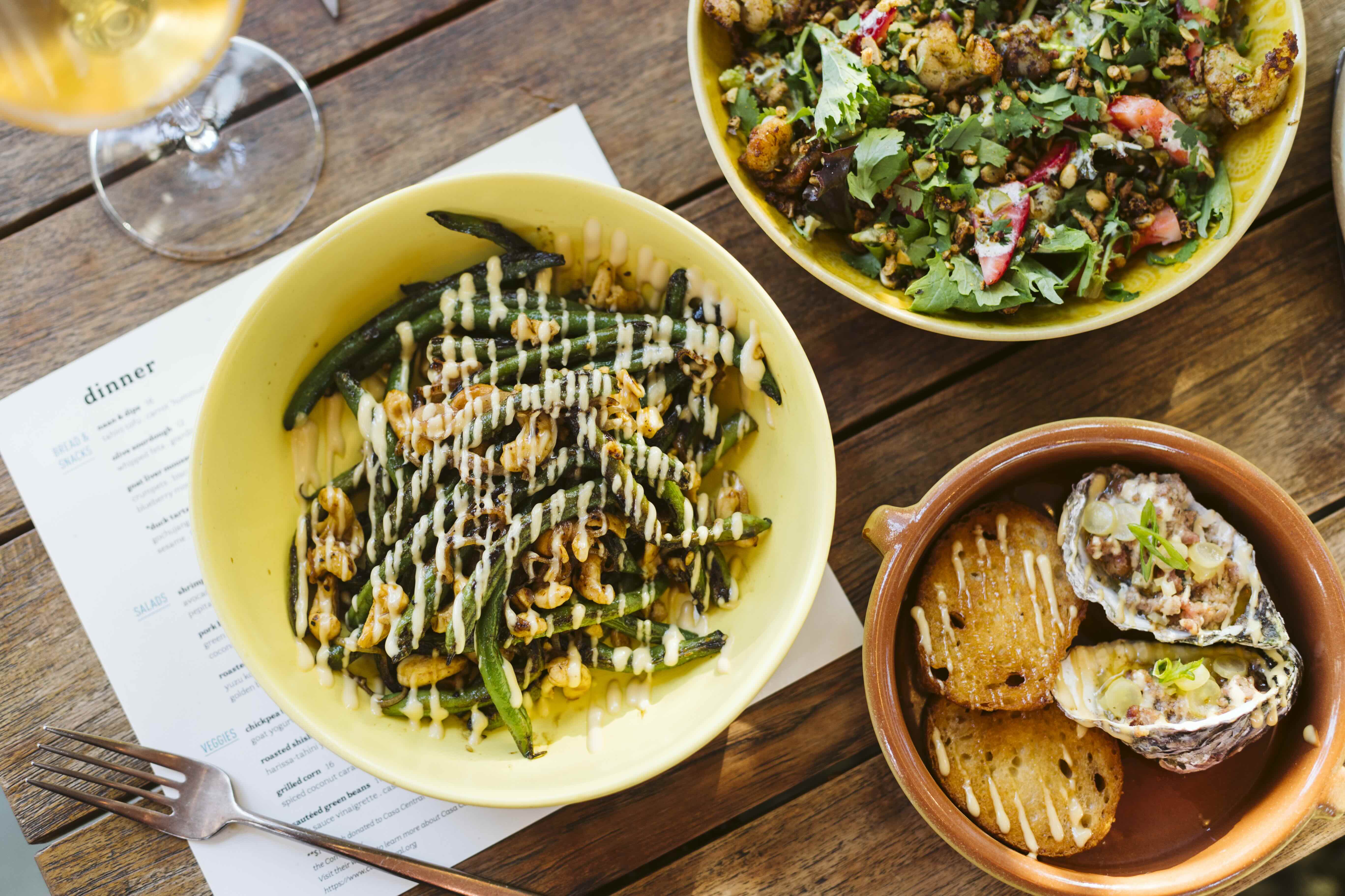 Yellow bowl of sautéed green beans with walnuts and creamy drizzle, brown bowl with grilled bread and oyster halves topped with garnish, and a bowl of mixed greens salad on a wooden table with a fork and a glass of white wine.