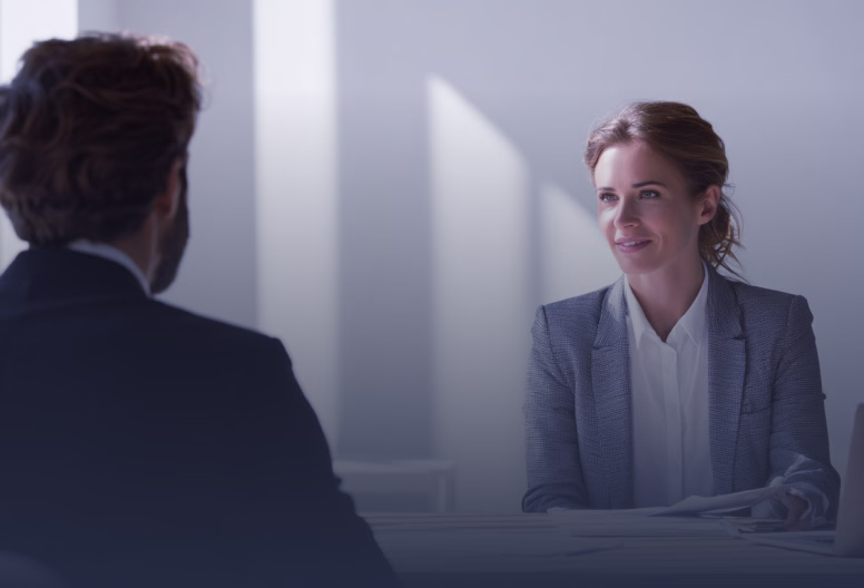 Woman in a grey blazer smiling and talking to a man in a dark suit across a table in a professional office setting.