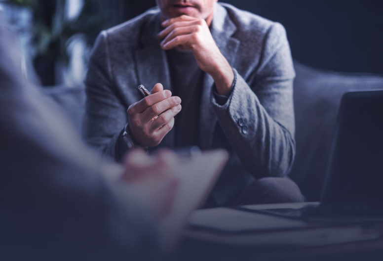 Man in grey blazer holding a pen, engaged in conversation during a meeting with a blurred person in foreground.