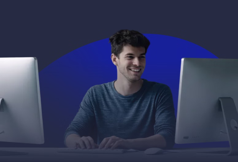 Smiling man in a blue long-sleeve shirt working at a desk with two computer monitors.
