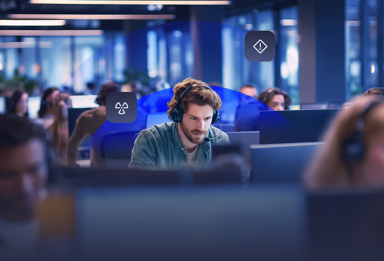 Man wearing headphones focused on computer screen in a busy modern office with digital hazard icons floating nearby.