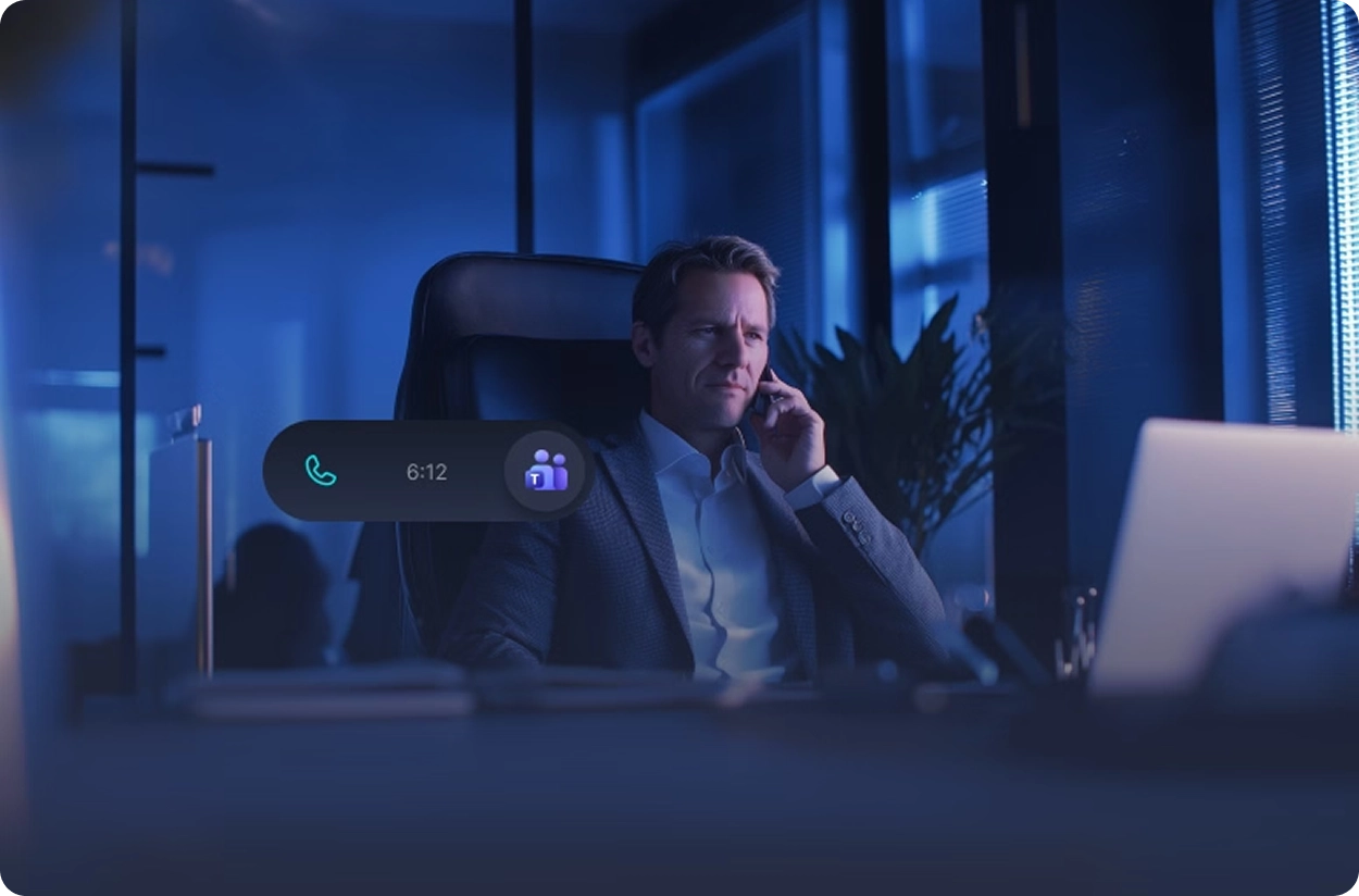 Man in suit sitting at desk in blue office talking on phone with Microsoft Teams call timer showing 6:12.