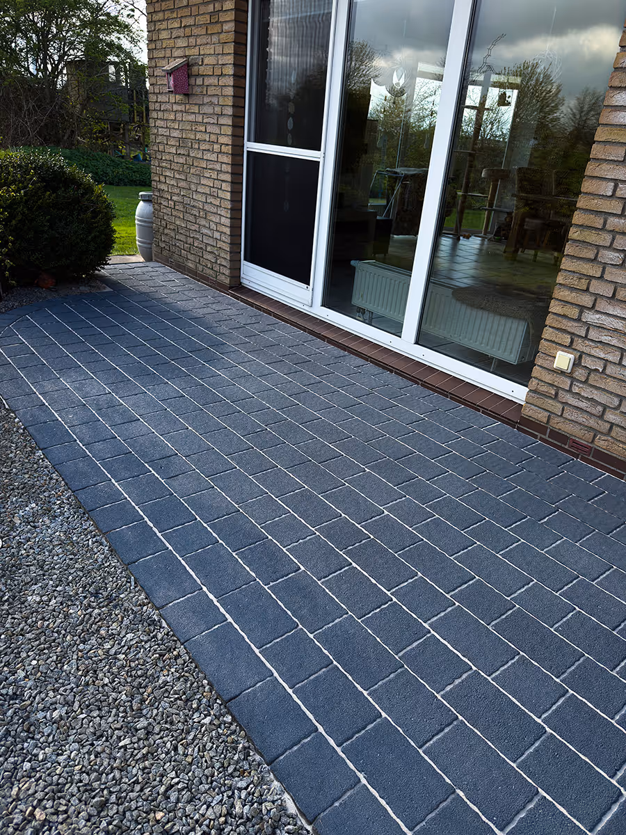 Terrassenbereich mit dunklen quadratischen Pflastersteinen neben einem Haus mit Glas-Schiebetür und Backsteinwand.