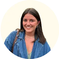 Smiling woman with straight brown hair wearing a denim jacket and layered necklaces against a neutral circular background.