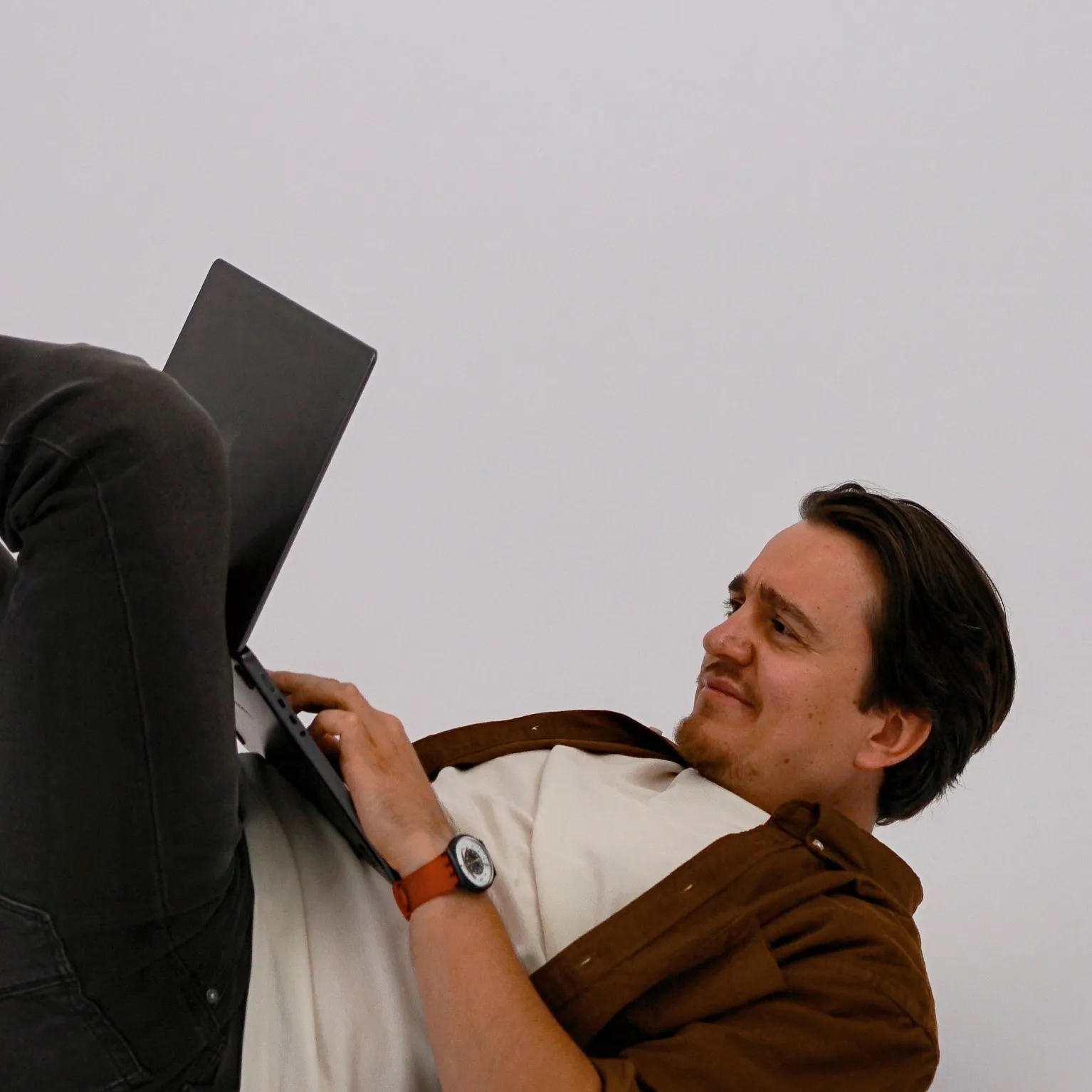 Man with a moustache and beard lying on his back against a plain white background, working on a laptop, wearing a brown shirt over a white t-shirt and a wristwatch with a brown strap.