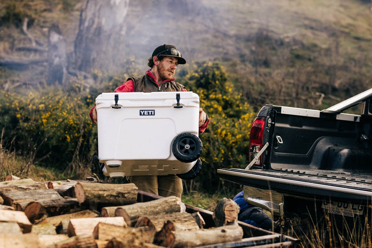 A man carrying a white YETI hard cooler to the back of a ute at camp