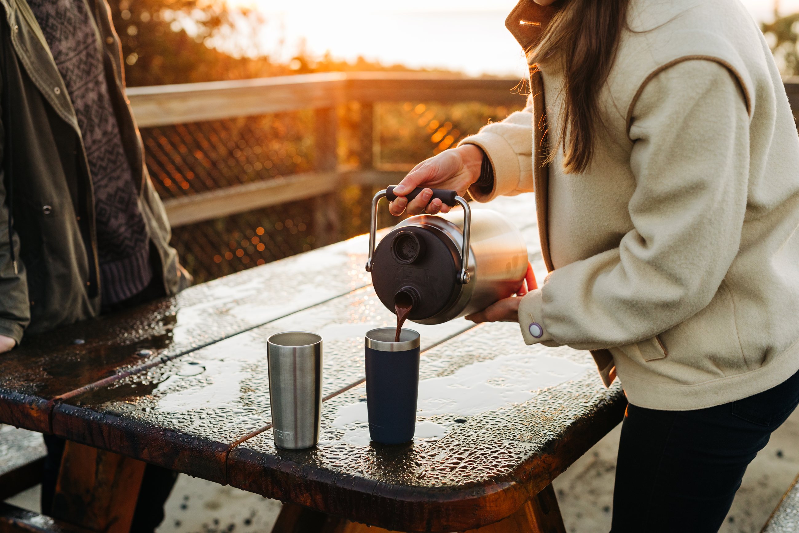 Coffee poured from a YETI jug into a tumbler on a wet picnic table at sunset