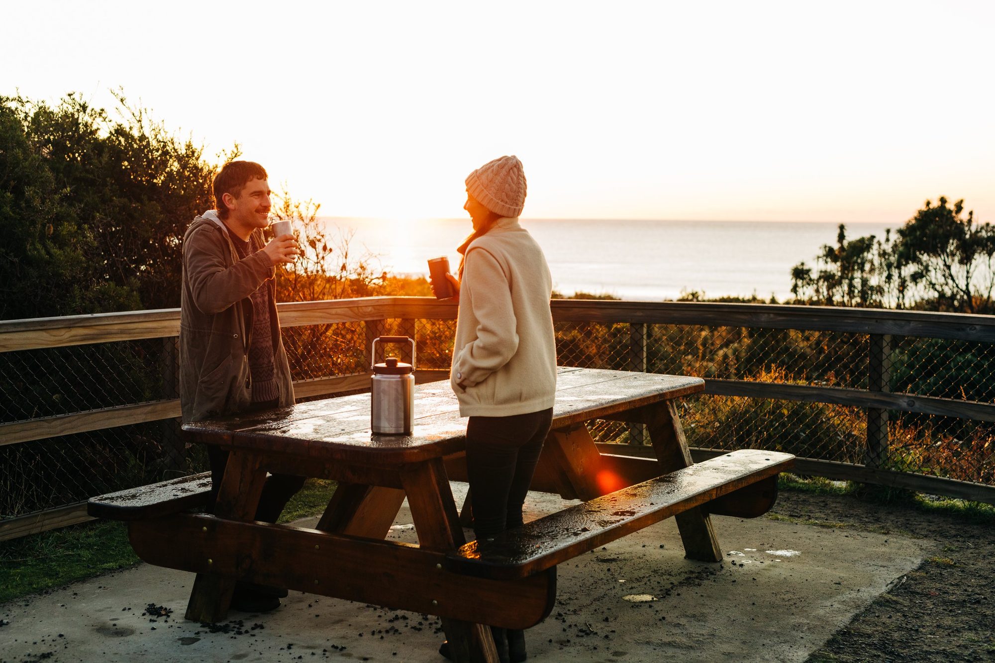 Two people sharing a hot drink from YETI drinkware at a picnic table at sunset