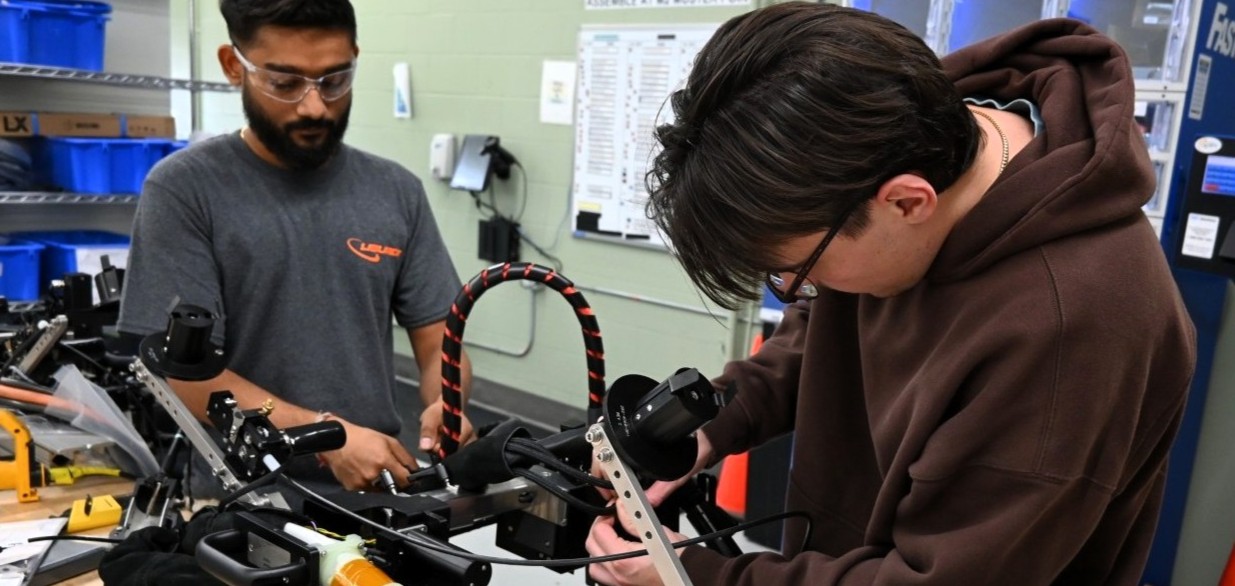 Two men work together on a mechanical device with tools in an industrial workshop setting.