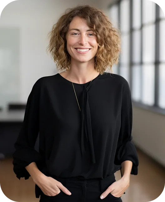 Smiling woman with curly hair wearing a black blouse and standing with hands in pockets in a bright room.