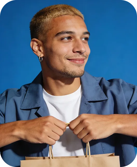 Young man with short blond hair and hoop earring smiling while holding a brown paper shopping bag against a blue background.